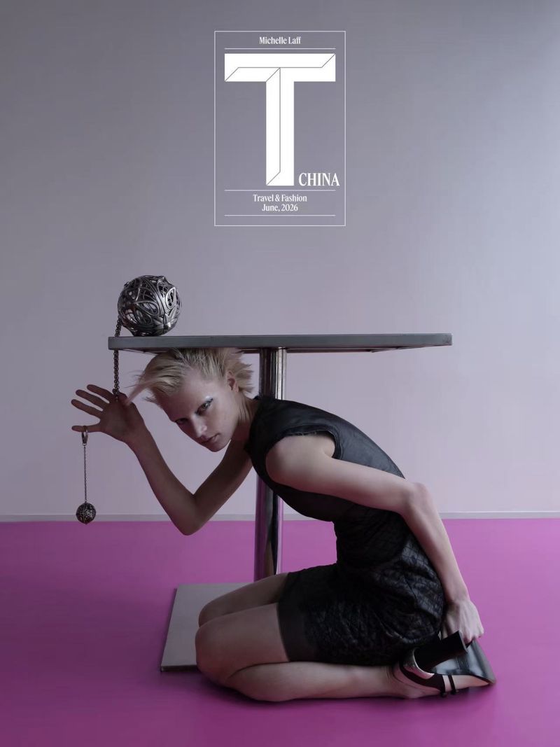 Michelle Laff wearing a black mini dress kneeling beneath a table holding a suspended sphere on a pink floor.