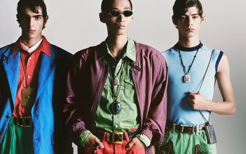 Three male models wearing colorful layered clothing and statement pendants against a neutral white backdrop.
