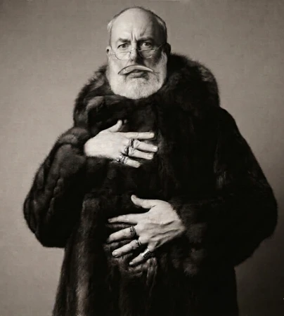 Elderly bearded man in a voluminous dark fur coat and glasses holding a small fish horizontally in his mouth.