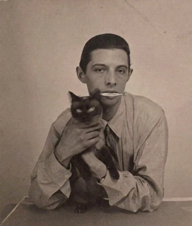 Vintage sepia portrait of a man in a collared shirt holding a fish in his mouth while holding a cat in his arms.