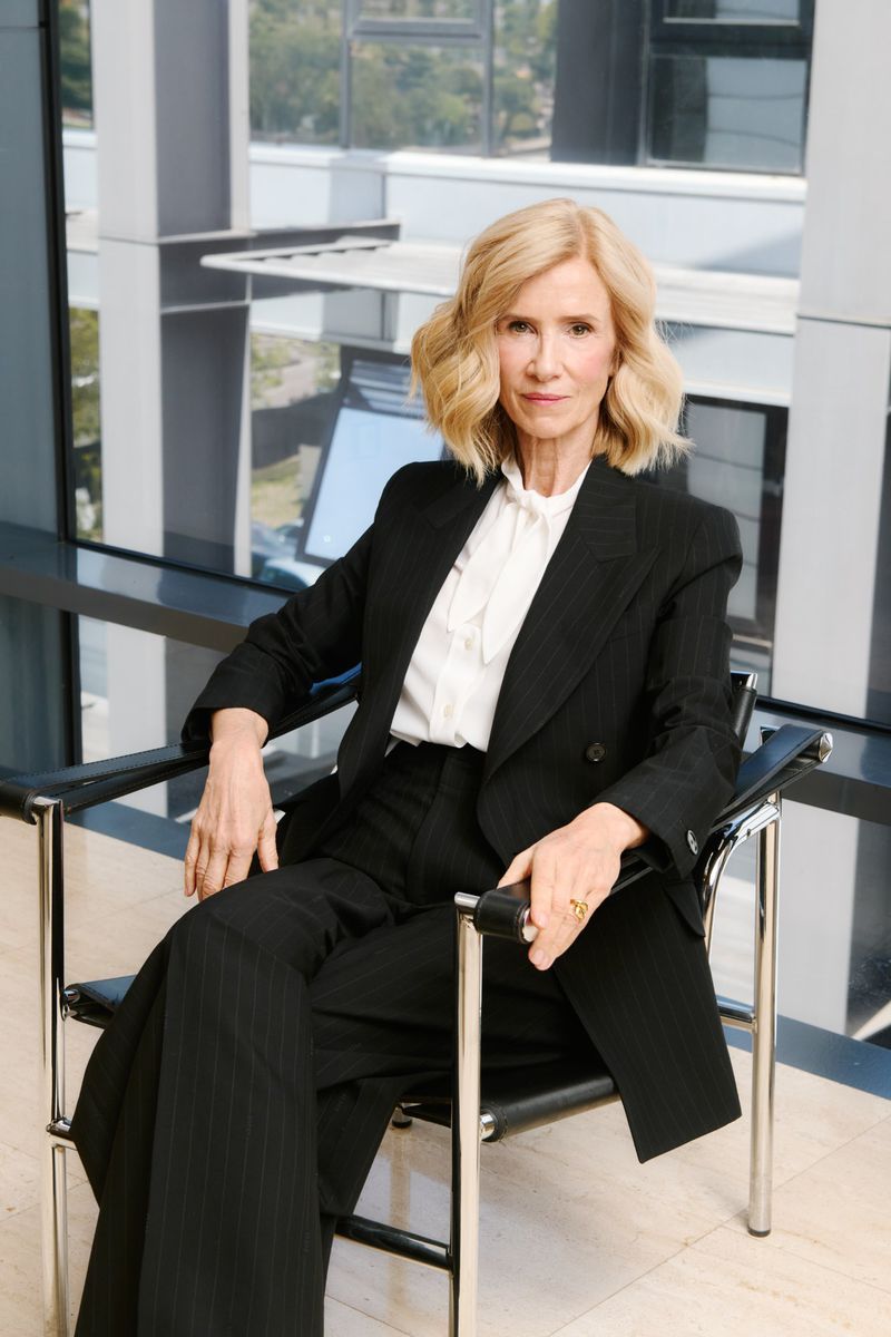 Professional portrait of a mature blonde woman in a black suit sitting in a modern chair in an office with natural light and neutral tones.