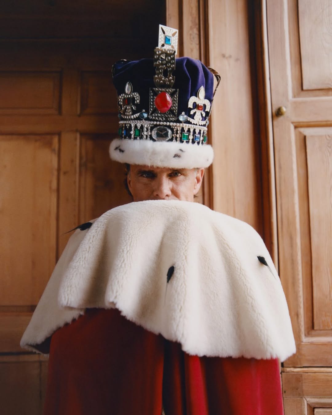 Walton Goggins wearing an ornate royal crown and pulling a white ermine fur cape over his lower face against wooden paneling.