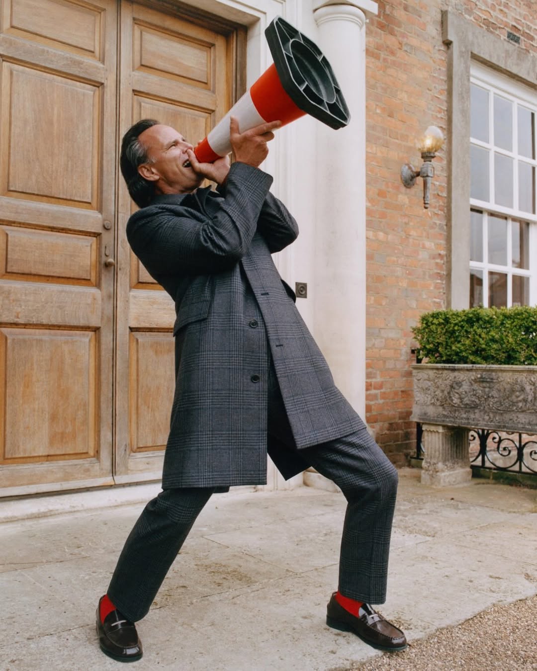 Actor Walton Goggins wearing a grey plaid coat and suit shouting into an orange traffic cone outside a stately brick entrance.