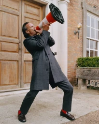Actor Walton Goggins wearing a grey plaid coat and suit shouting into an orange traffic cone outside a stately brick entrance.