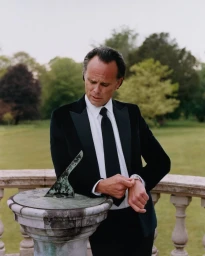 Walton Goggins in a black tuxedo and tie checking his watch beside an antique stone sundial on an outdoor terrace.