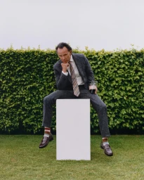 Walton Goggins seated on a white plinth holding a drink in front of a green hedge, wearing a grey suit and striped tie.