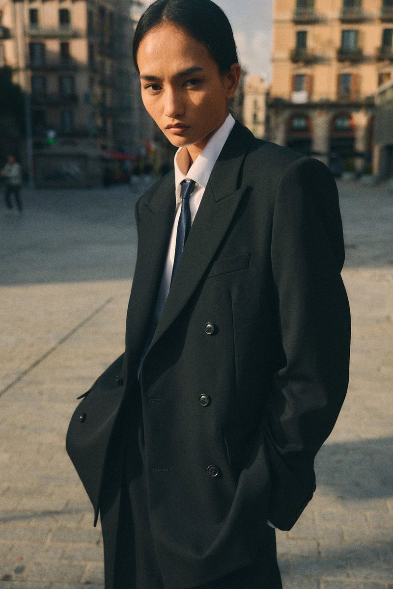 Model Mona Bhujel wearing an oversized black double-breasted blazer and tie in an outdoor urban square.