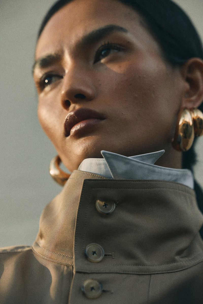 Model Mona Bhujel wearing a high-collar beige coat and sculptural gold earrings in warm sunlight.