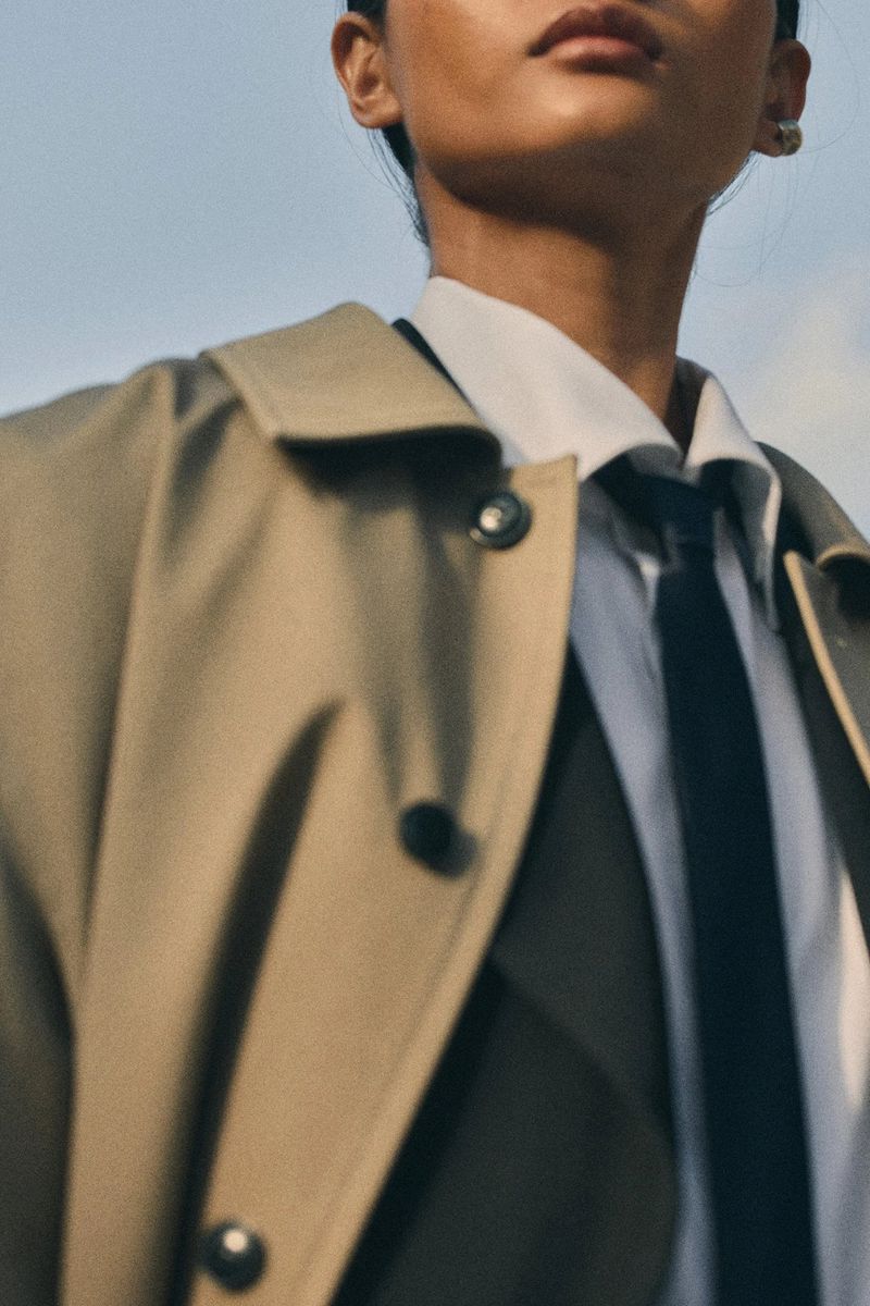 Low-angle close-up of model Mona Bhujel wearing a beige trench coat over a white collared shirt and black tie against an open sky.