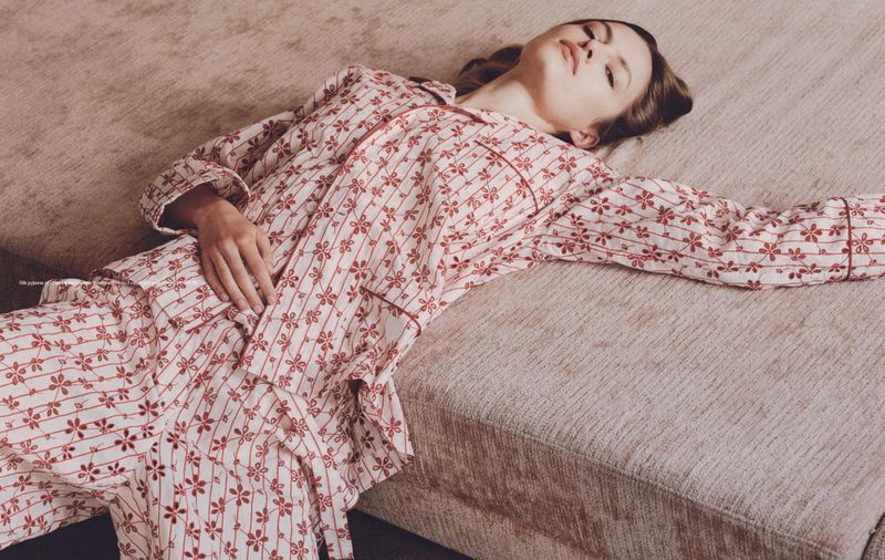 A young adult female lies on a light beige sofa in red-and-white floral pajamas, relaxed and looking upwards in a styled editorial fashion shot.