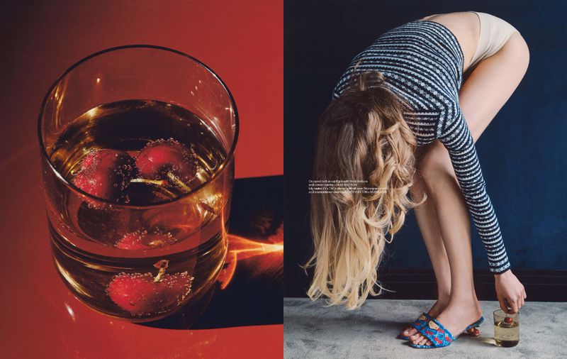 Woman bends at waist, holding glass with cherries. Second glass with cherries sits on red table. Navy wall background.