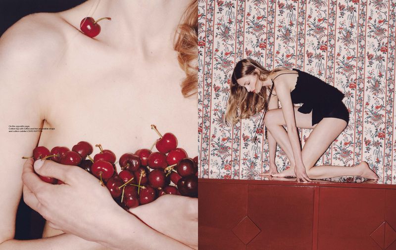 Conceptual fashion shot: fair-skinned woman covered with cherries, posing in black lingerie, floral wallpaper, red sideboard.