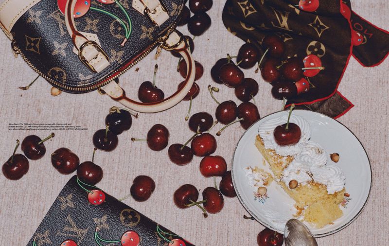 Overhead shot of designer purse and scarf scattered with cherries and piece of cake topped with whipped cream and cherry, indoor setting.