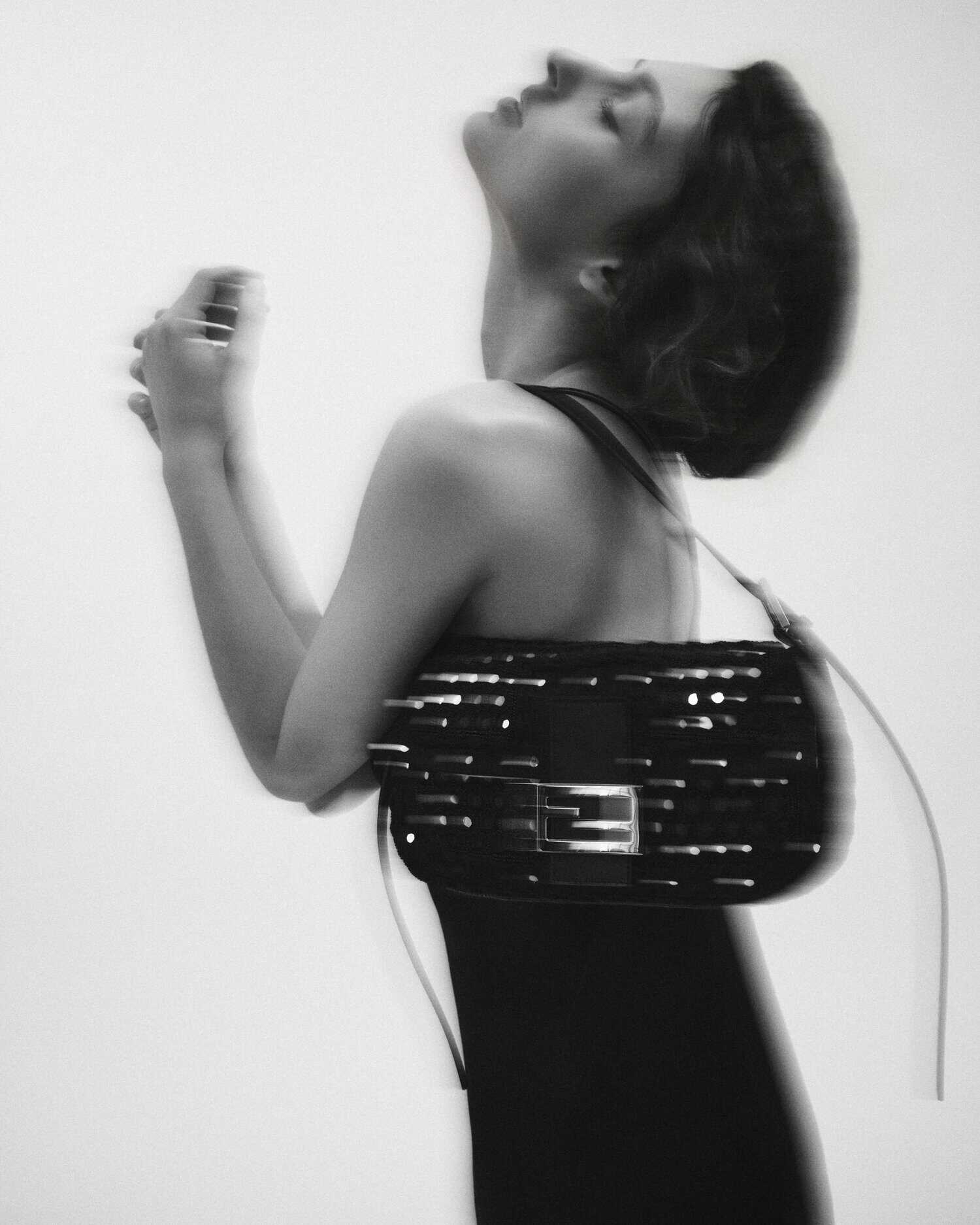 Monochrome portrait of a woman in a black dress posing with an embellished black Fendi Baguette shoulder bag on a white backdrop.