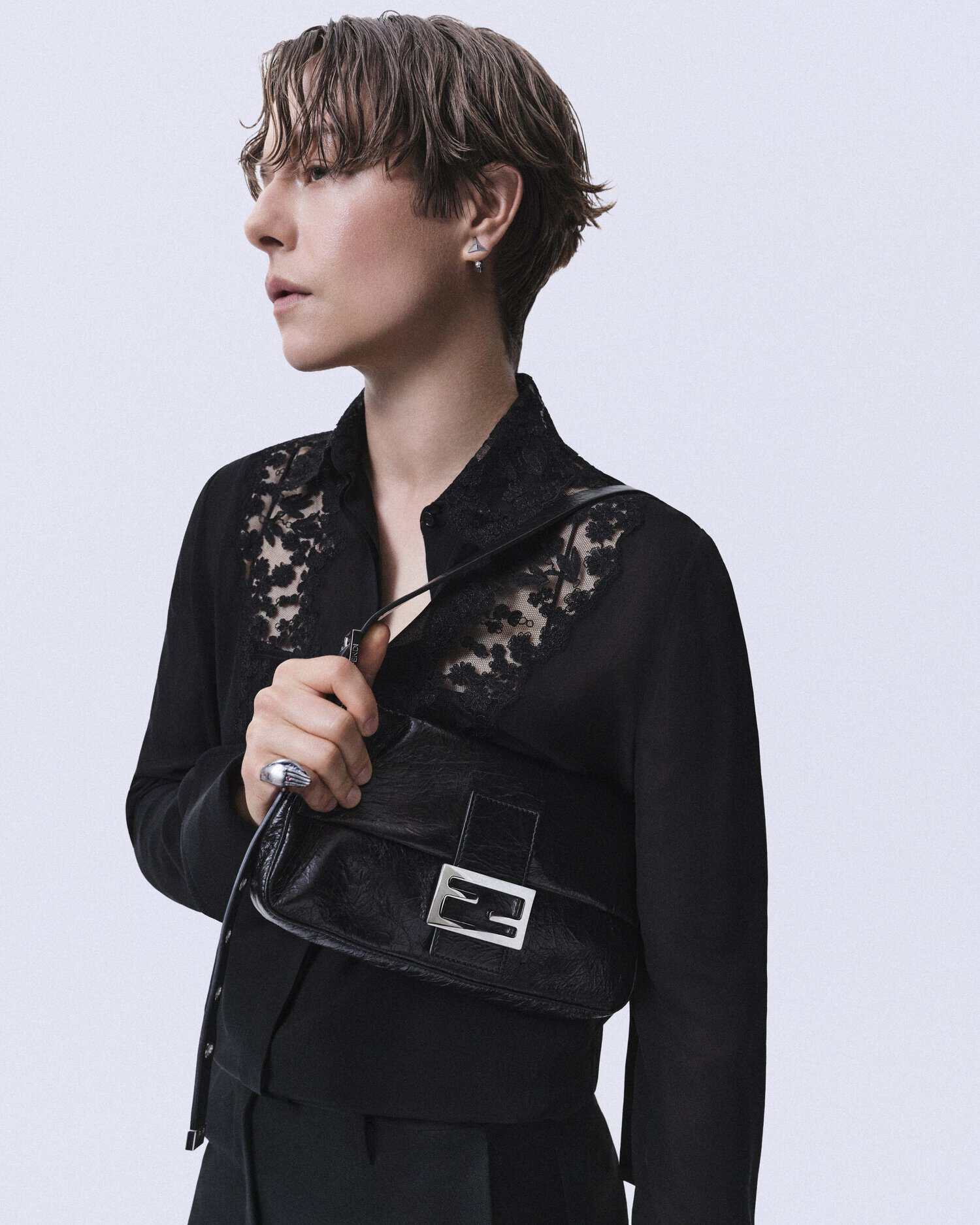 Emma D'Arcy holding a black leather Fendi Baguette bag while wearing a sheer lace-panel black shirt against a light studio backdrop.