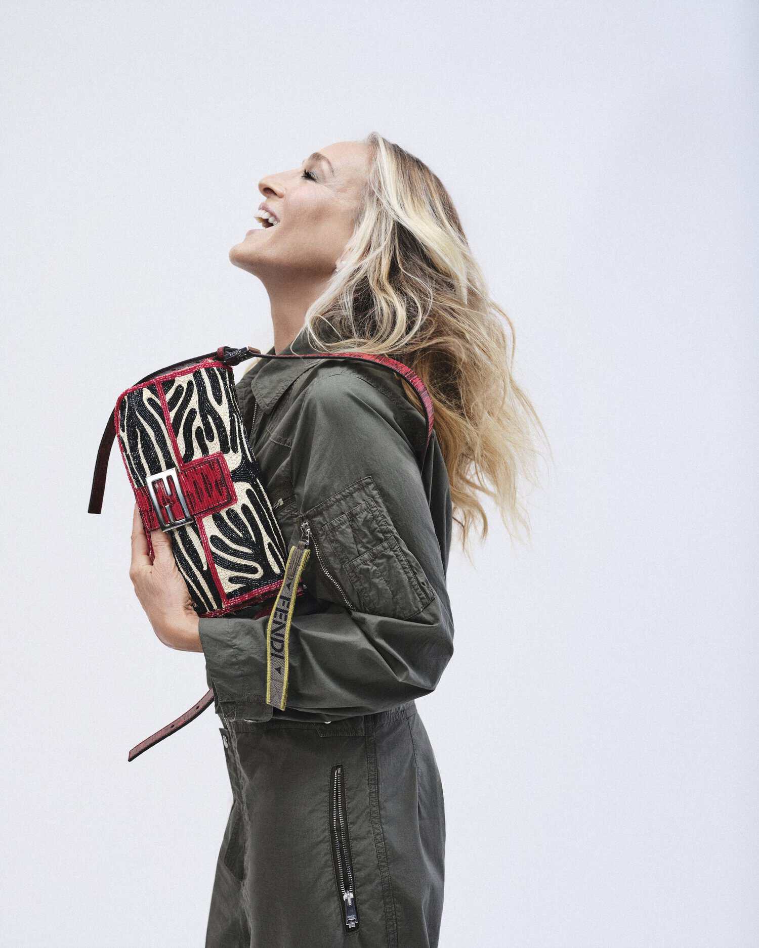 Sarah Jessica Parker wearing an olive green jumpsuit holding a patterned Fendi Baguette shoulder bag against a white background.