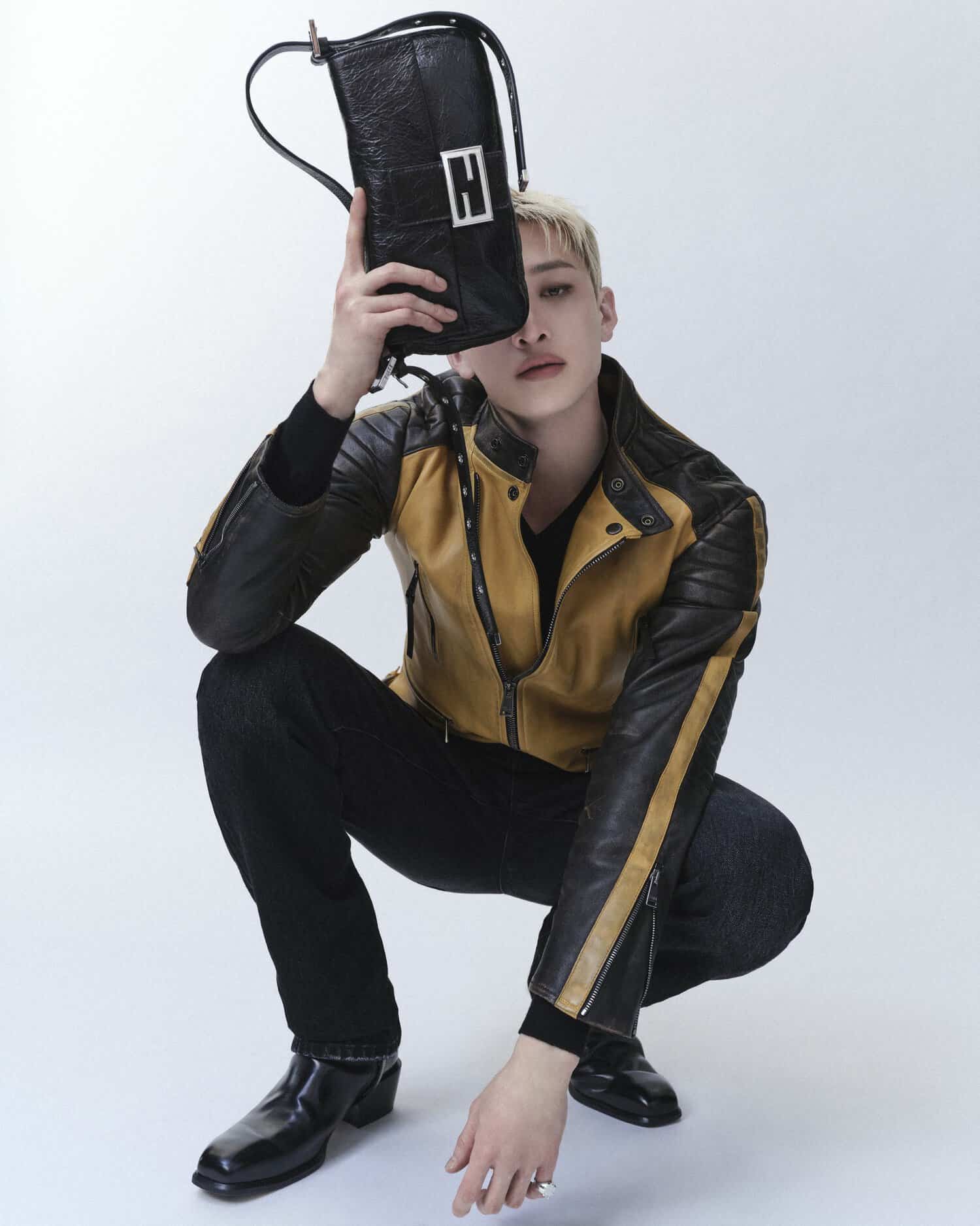 Bang Chan crouching in a yellow and black leather jacket holding a black leather Fendi Baguette bag over his face in a studio.