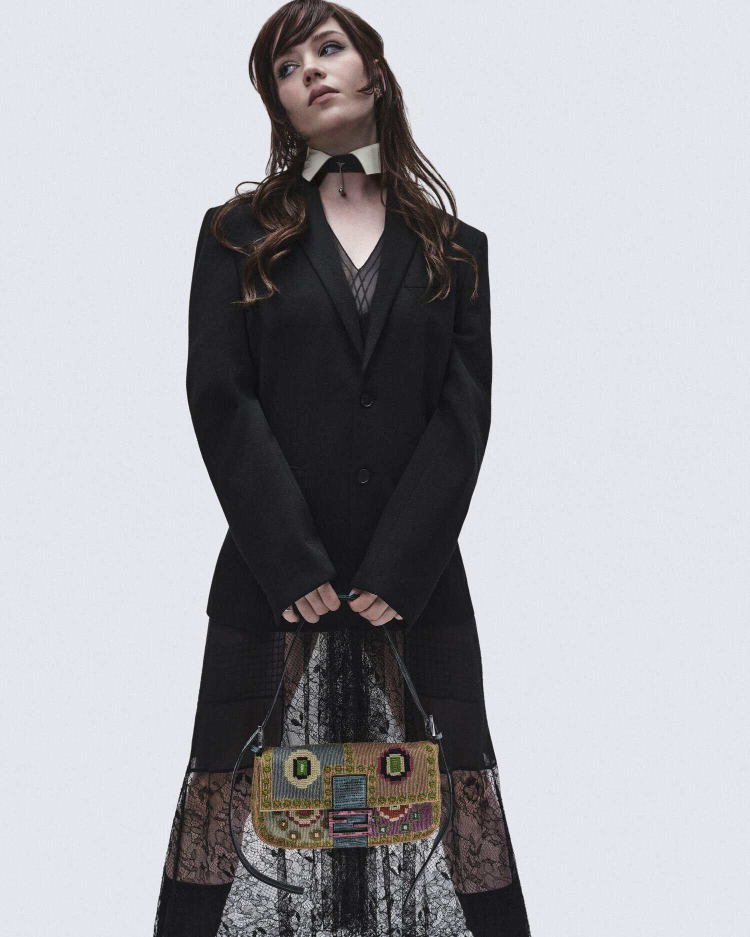 Sophie Thatcher posing in a black blazer and lace skirt holding an embroidered Fendi Baguette bag against a light studio backdrop.