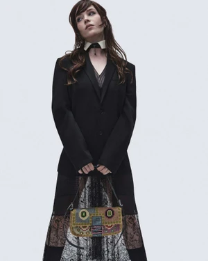 Sophie Thatcher posing in a black blazer and lace skirt holding an embroidered Fendi Baguette bag against a light studio backdrop.