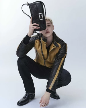 Bang Chan crouching in a yellow and black leather jacket holding a black leather Fendi Baguette bag over his face in a studio.