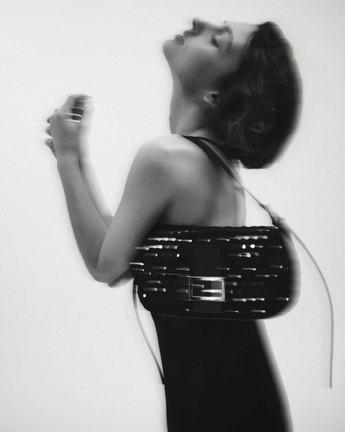 Monochrome portrait of a woman in a black dress posing with an embellished black Fendi Baguette shoulder bag on a white backdrop.
