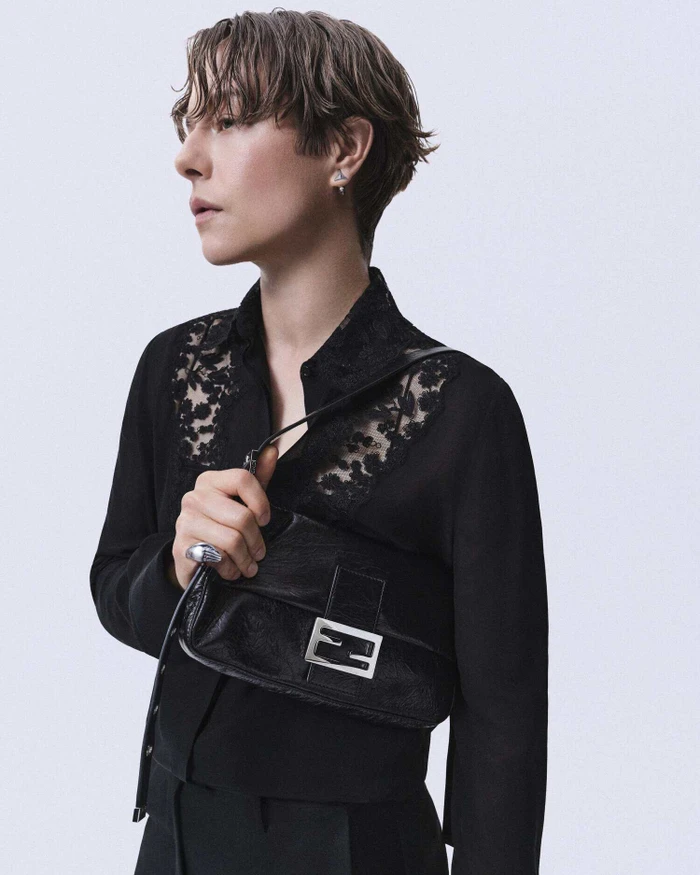 Emma D'Arcy holding a black leather Fendi Baguette bag while wearing a sheer lace-panel black shirt against a light studio backdrop.