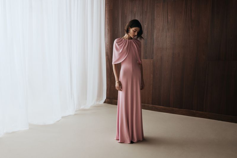 Model in a draped pink gown standing indoors against a dark wood wall and sheer white curtains