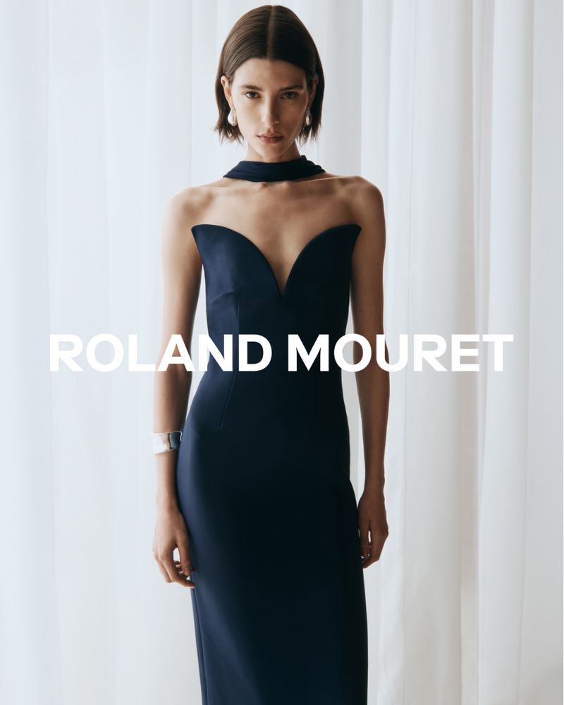 Model wearing a dark strapless evening dress with a neck scarf standing before white sheer curtains.
