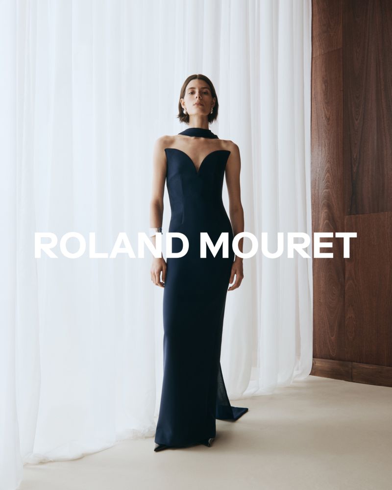 Model Erika Barletta wearing a dark navy strapless sculpted evening gown with a high neck scarf detail against sheer white curtains.