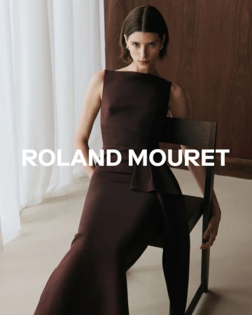 Model seated on a dark wooden chair wearing a sleek dark brown sleeveless midi dress against a white curtain background.