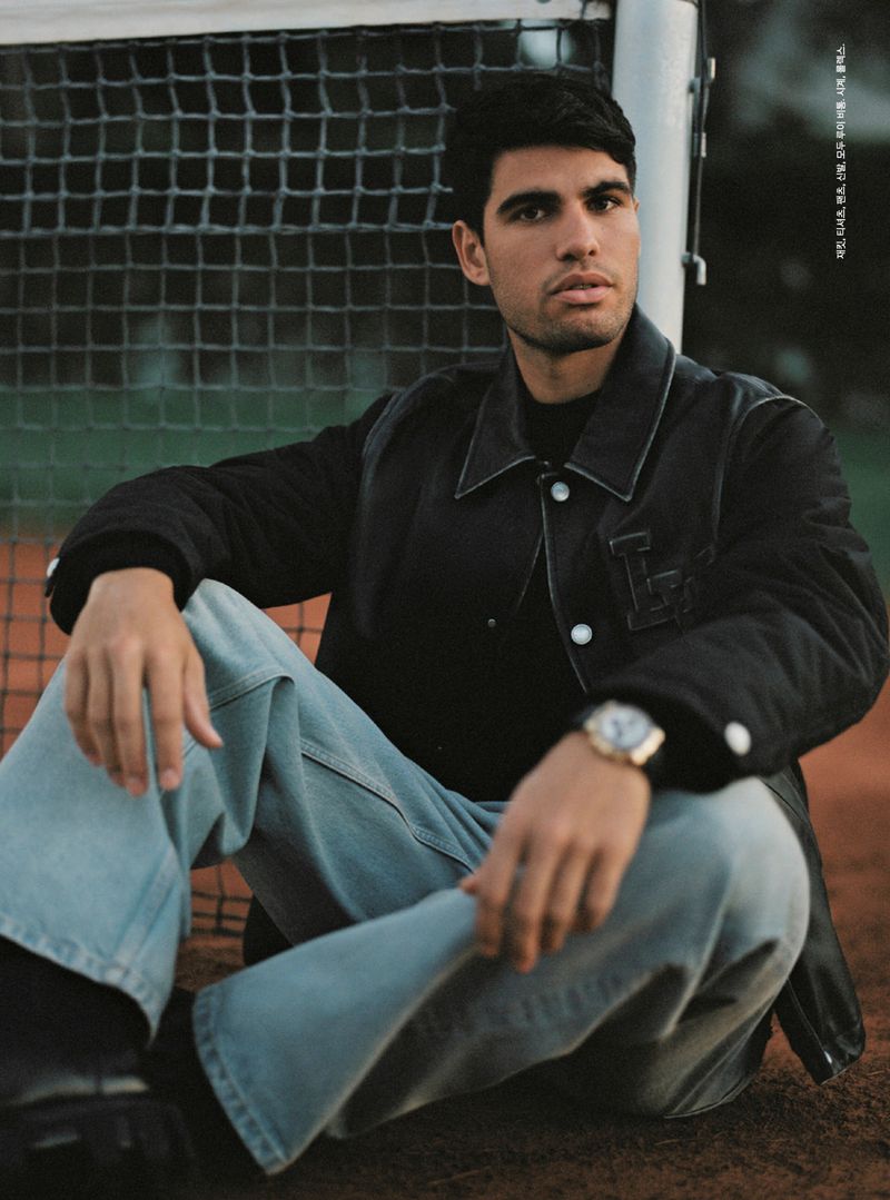 Male model in a black quilted jacket and light blue jeans sitting on a red clay surface with a blurred netting background.