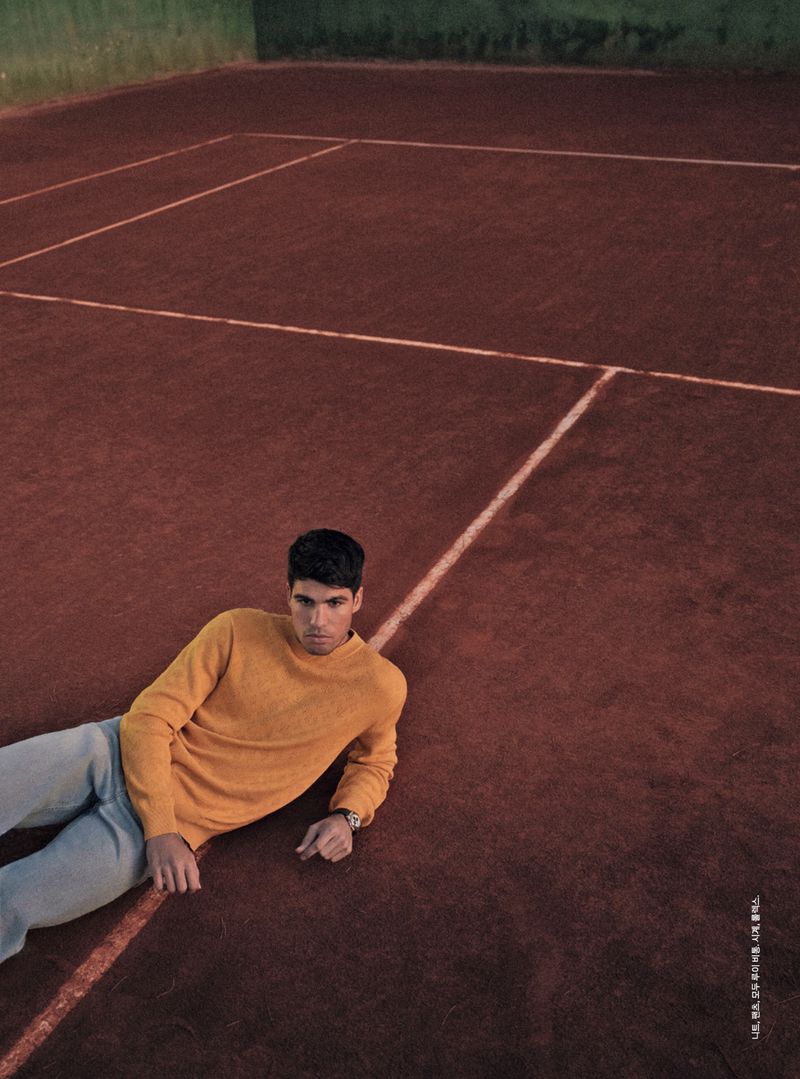 Man in yellow sweater and jeans lies on a red clay tennis court with white lines. He is wearing a watch and looking at the camera.