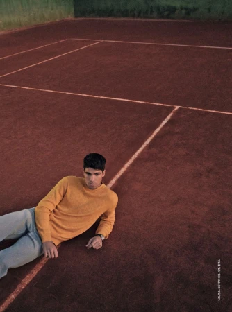 Man in yellow sweater and jeans lies on a red clay tennis court with white lines. He is wearing a watch and looking at the camera.