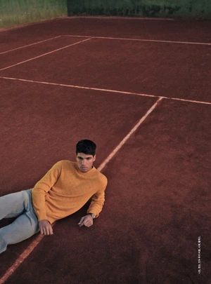 Carlos Alcaraz lying on a red clay tennis court wearing a mustard yellow knit sweater and light-wash trousers.