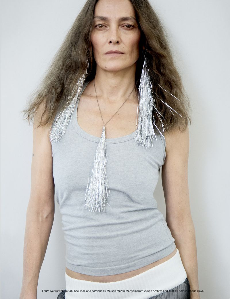 Adult female with long brown hair wearing a gray tank top, silver fringe earrings and necklace stands in front of a white wall.