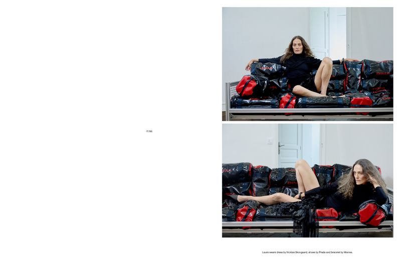 A woman in a black turtleneck and skirt sits on a platform covered in red and black duffel bags in a brightly lit indoor scene.