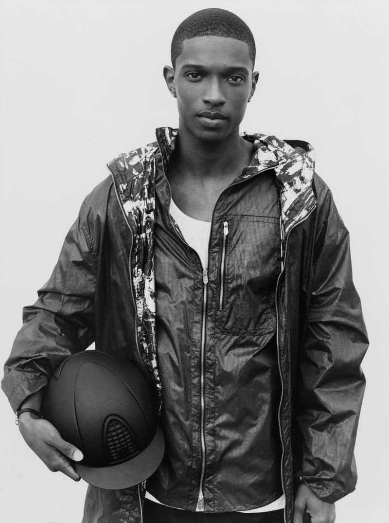 Portrait of an adult male wearing a jacket and holding a black equestrian helmet against a plain backdrop in a studio setting.