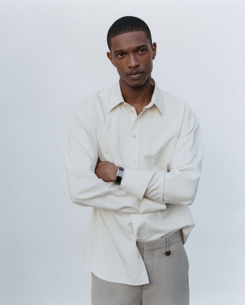 Portrait of adult male with crossed arms wearing a striped button-down shirt and khaki trousers against a solid neutral background.