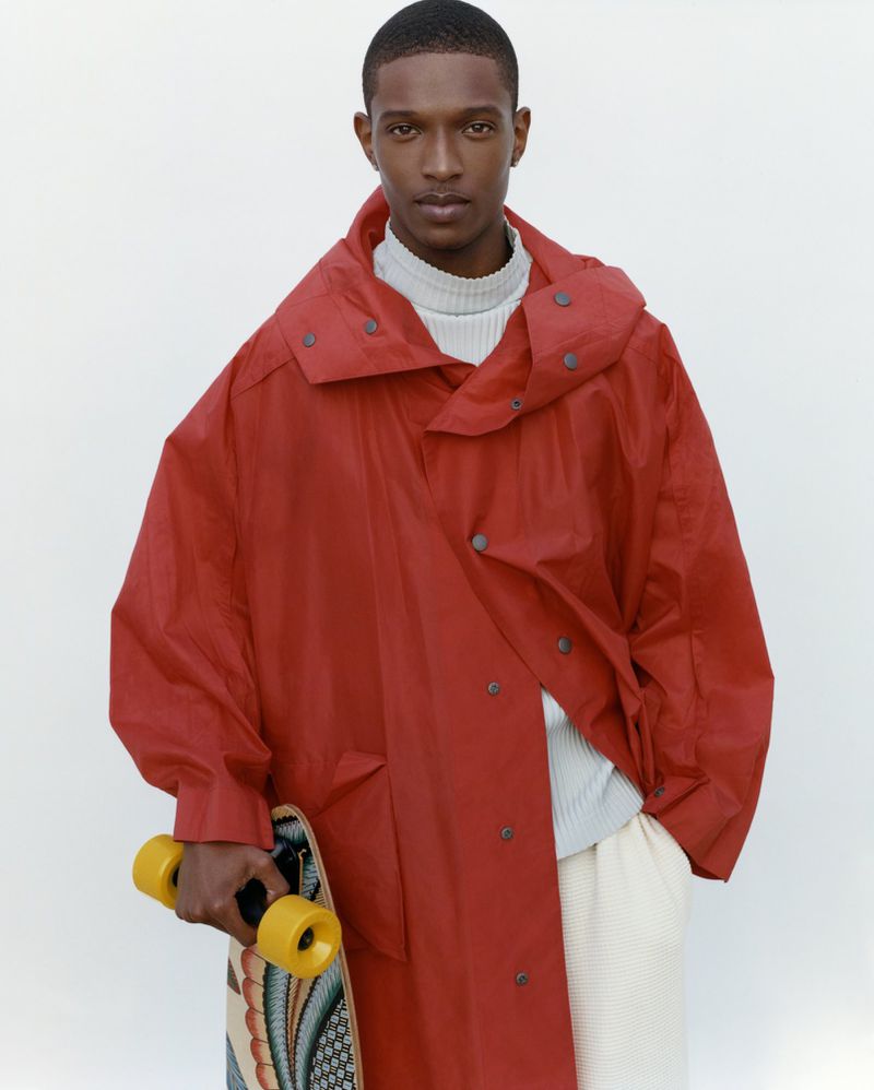 An adult Black male in a red coat and light trousers holds a skateboard against a plain background. The light is bright and diffused.