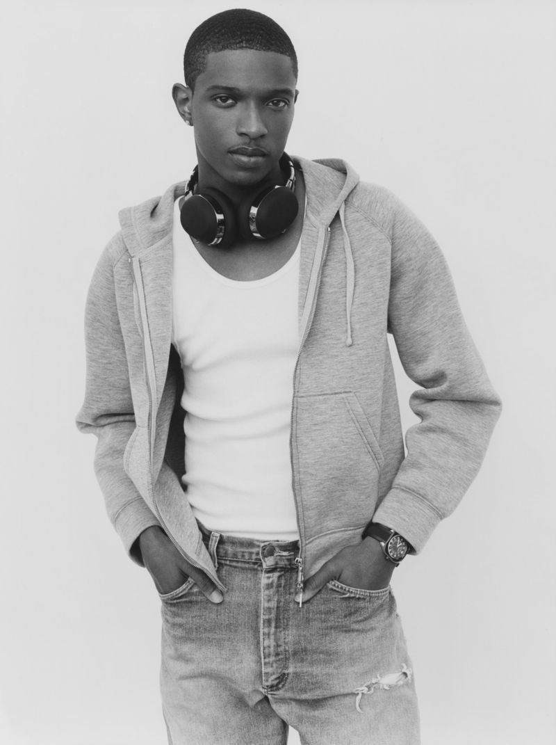 Model Malik Anderson wearing a grey zip-up hoodie, white tank top, and over-ear headphones around his neck against a studio backdrop.
