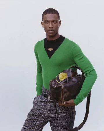 An African-American man stands indoors in a studio setting, wearing a green v-neck sweater, tweed pants, and carrying a brown leather duffel bag.