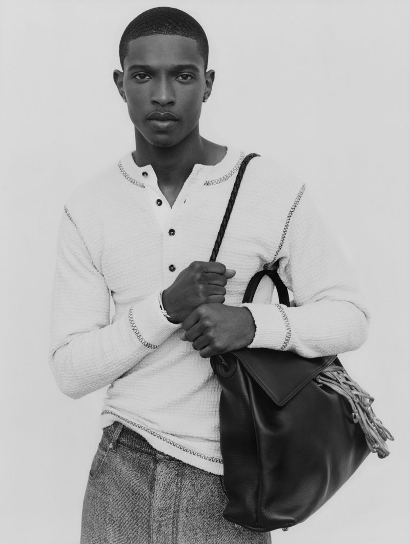 Black and white portrait of a dark-skinned man wearing a white waffle-knit Henley shirt and grey tweed pants, holding a black leather backpack.
