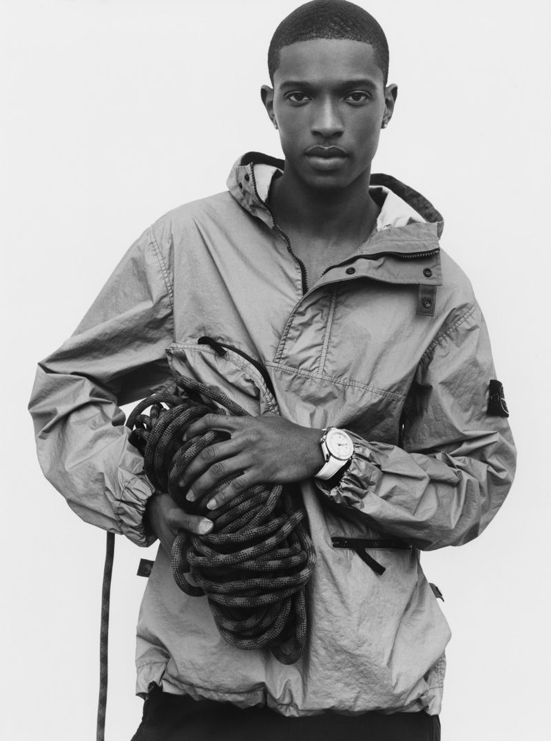 Portrait of a young man holding a rope, wearing a hooded jacket and watch, against a bright background, in a fashion editorial style.