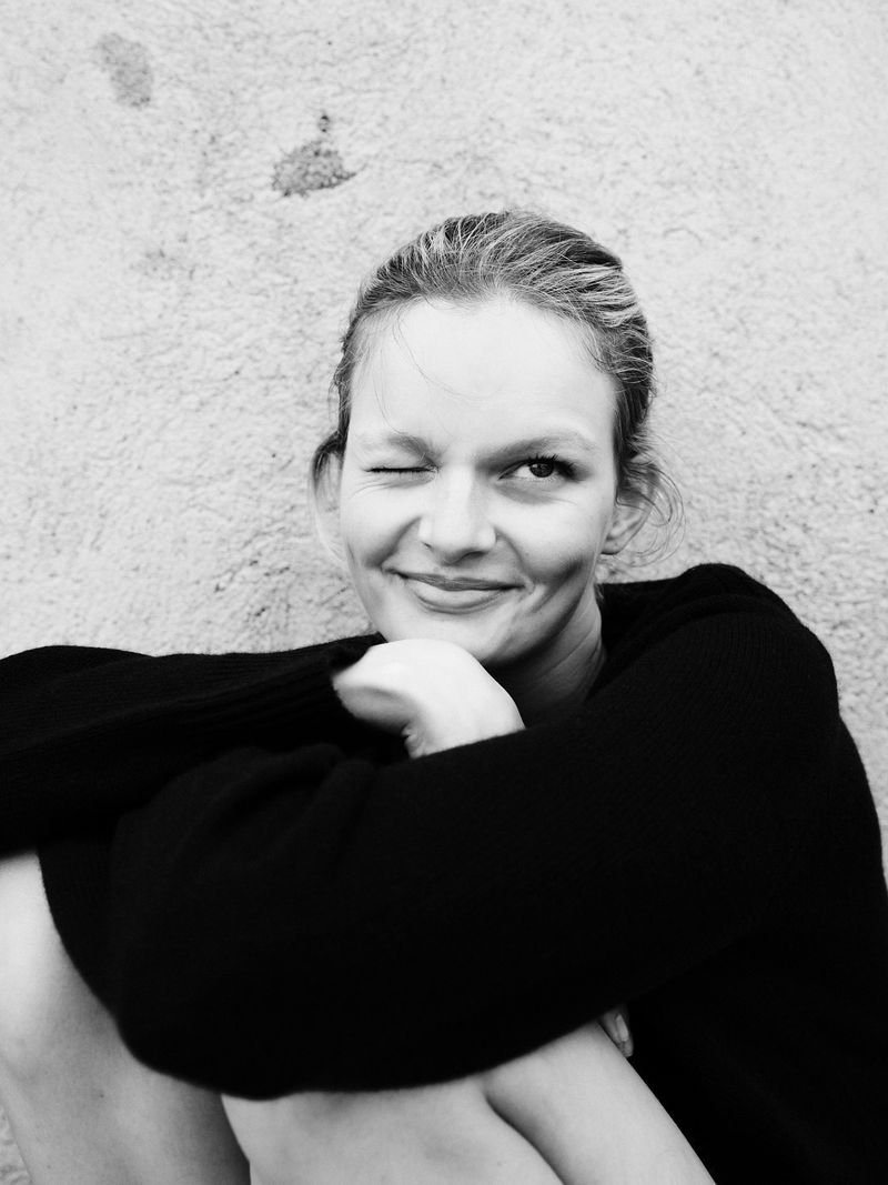 Model Bo Exters playfully winking and smiling while wearing a black knit sweater against a textured wall.