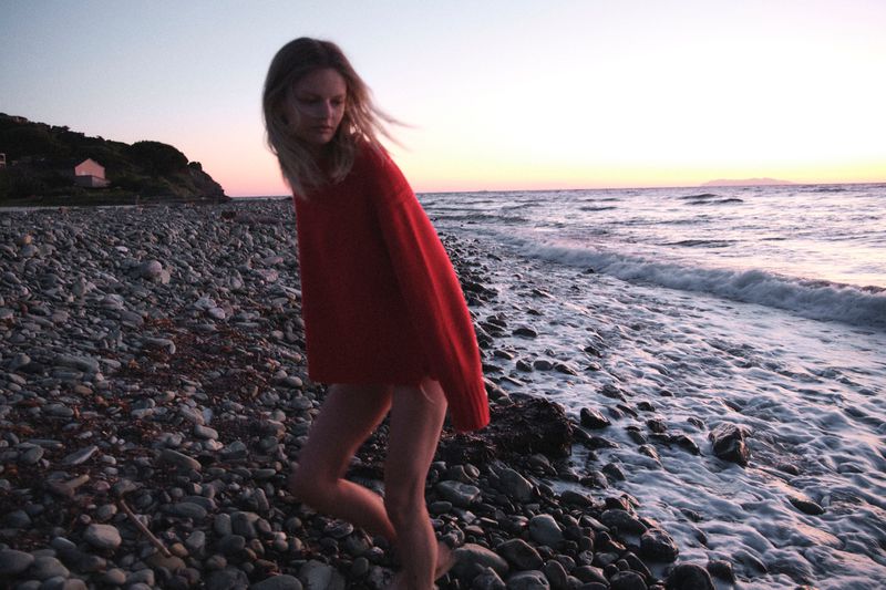 Model Bo Exters walking along a pebble beach at sunset wearing a bright red oversized knit sweater.