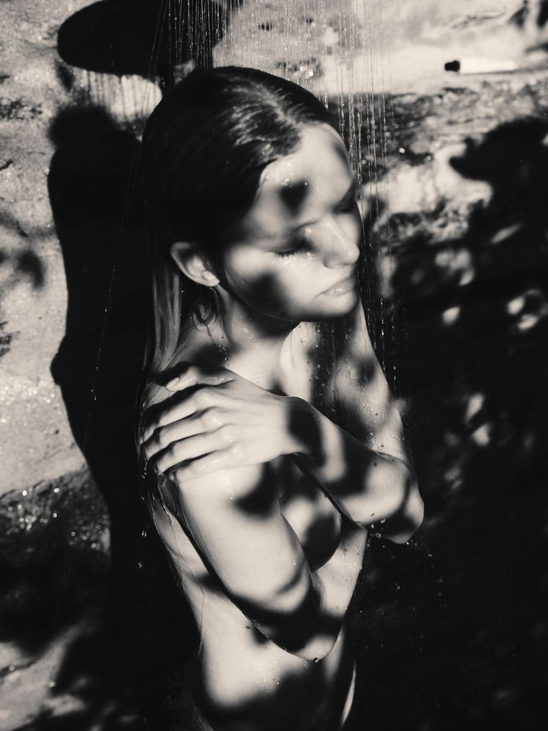 Black and white portrait of model Bo Exters under an outdoor shower with dappled foliage shadows on her skin.