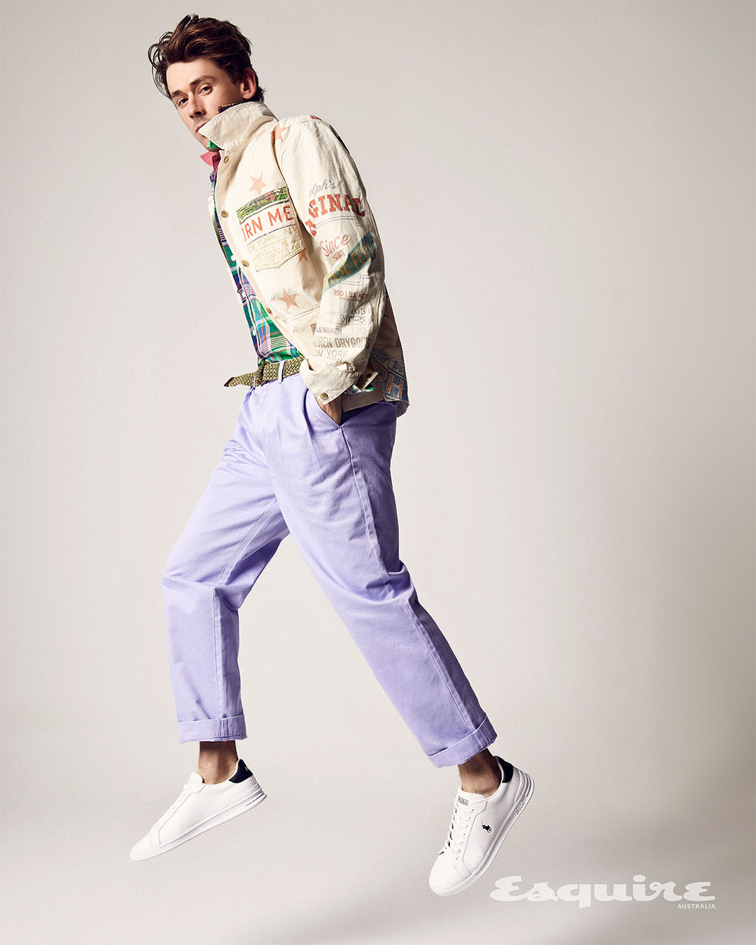 Full shot of a young adult male model mid-air in a light patterned jacket, colorful shirt, lilac pants, and white sneakers against a white background.