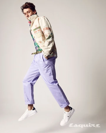 Full shot of a young adult male model mid-air in a light patterned jacket, colorful shirt, lilac pants, and white sneakers against a white background.