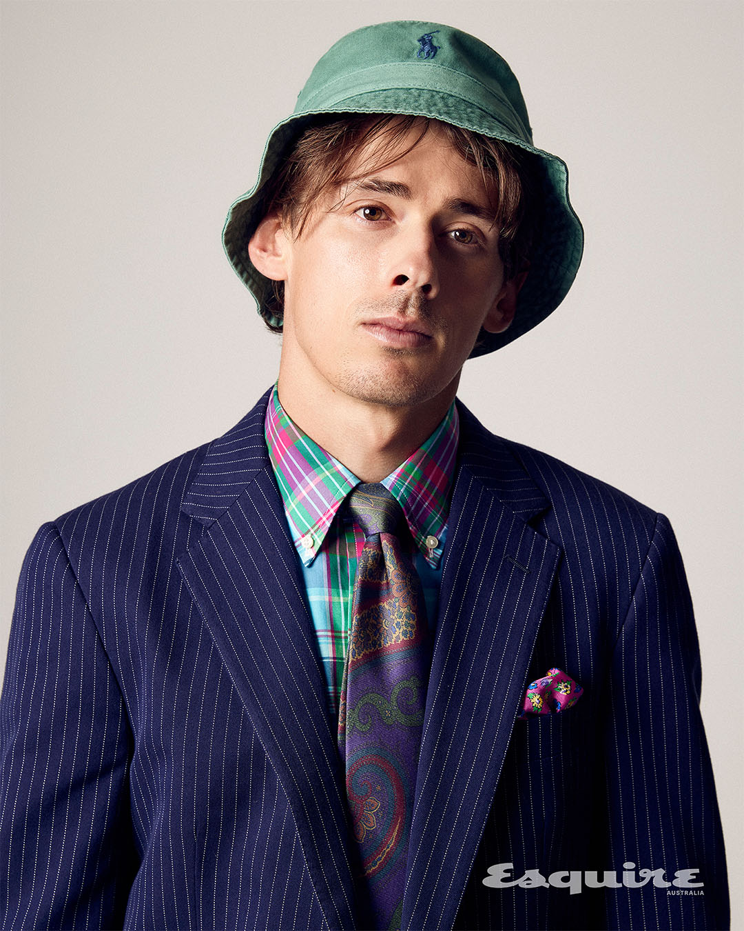 Stylish man in a pinstripe suit, patterned tie, and mint green bucket hat posing in a studio, looking directly at the camera.