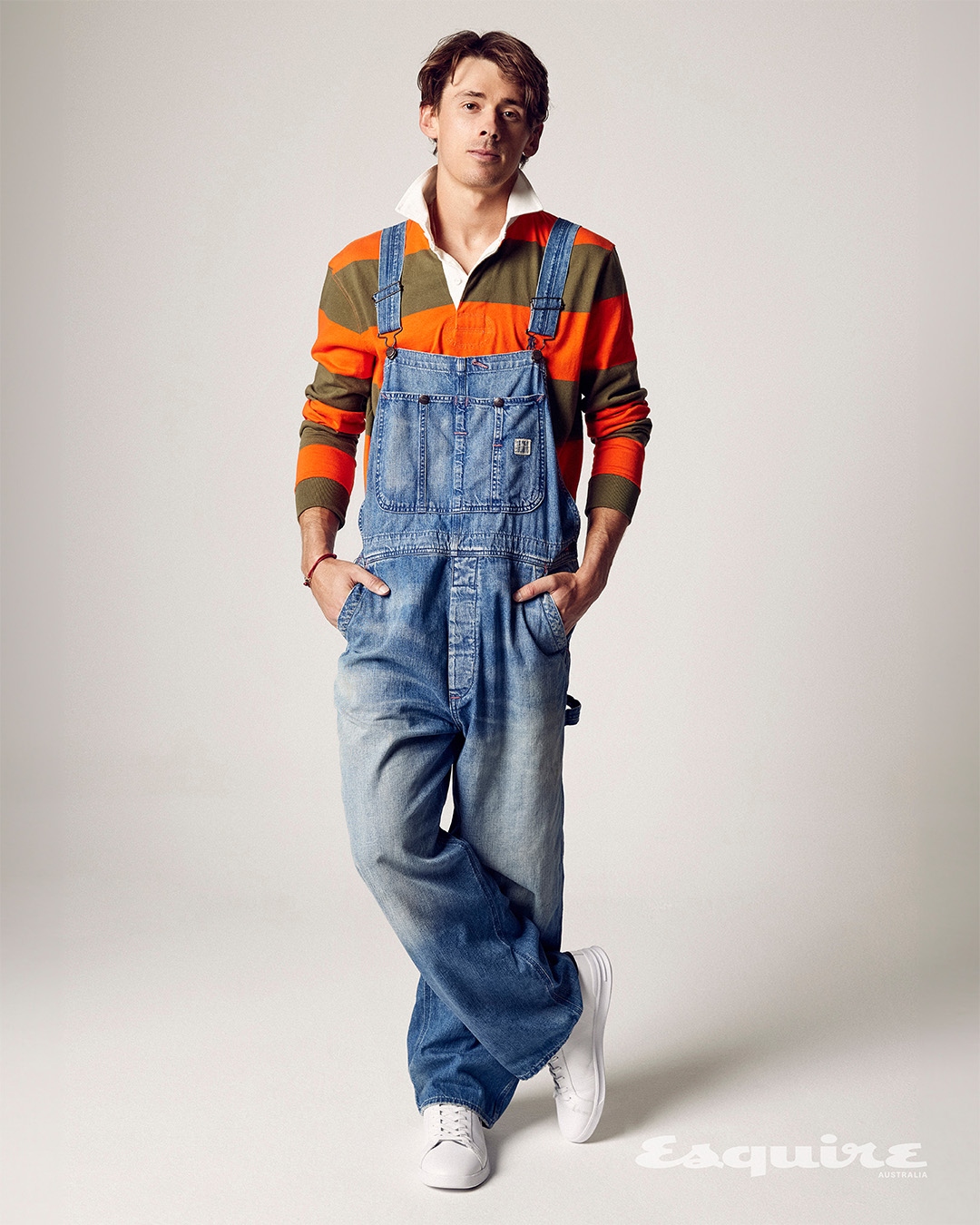 Full-body shot of a male model in a striped shirt and denim overalls posing indoors against a plain white background in a photo studio.